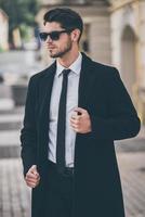 élégant et beau. beau jeune homme bien habillé en lunettes de soleil ajustant son manteau et regardant ailleurs tout en se tenant à l'extérieur photo