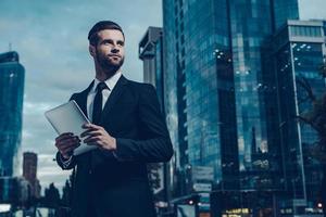 c'est ma ville. image de nuit d'un jeune homme confiant en costume complet tenant une tablette numérique et regardant ailleurs tout en se tenant à l'extérieur avec le paysage urbain en arrière-plan photo