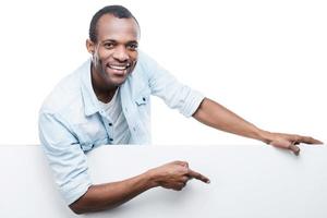 bel espace de copie de pointage. joyeux homme africain en chemise bleue se penchant sur l'espace de copie et le pointant en se tenant debout sur fond blanc photo