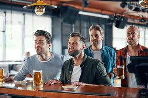 acclamant les jeunes hommes en tenue décontractée regardant un match de sport et dégustant une bière assis dans le pub photo