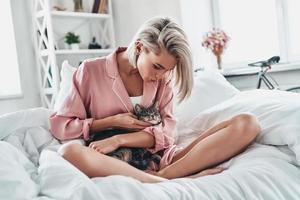 sa petite amie. jolie jeune femme jouant avec son chat et souriant assis dans son lit à la maison photo
