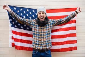 patriote excité de style américain. excité jeune homme tenant un drapeau américain debout contre un mur de briques photo