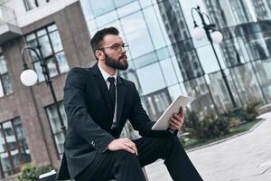 envisager la prochaine étape. beau jeune homme en costume complet à l'aide d'une tablette numérique tout en étant assis à l'extérieur photo