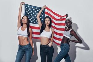 charmants patriotes. trois jolies jeunes femmes tenant un drapeau américain et regardant la caméra en se tenant debout sur fond gris photo