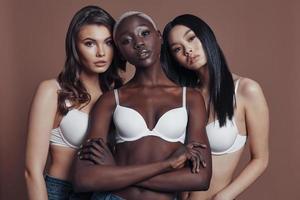 trop beau pour être vrai. trois jolies jeunes femmes regardant la caméra en se tenant debout sur fond marron photo