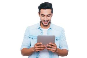 examinant son tout nouveau gadget. beau jeune homme indien tenant une tablette numérique et le regardant avec le sourire en se tenant debout sur fond blanc photo