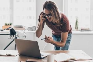 occupé à travailler au bureau. jeune homme concentré aux cheveux longs regardant son ordinateur portable et pointant le moniteur avec un stylo tout en se tenant près de son lieu de travail au bureau photo