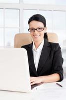 femme d'affaires au travail. confiante jeune femme d'affaires utilisant un ordinateur et souriante assise sur son lieu de travail photo