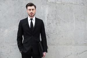 confiant et autoritaire. beau jeune homme tenant une mallette debout à l'extérieur et contre le mur de béton photo