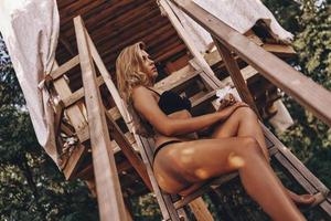 trop beau pour être vrai. jolie jeune femme en bikini noir regardant loin tout en étant assis sur les marches en bois de la cabane de plage photo