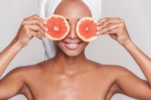 station thermale naturelle. belle jeune femme torse nu afro-américaine tenant des morceaux d'orange devant ses yeux en se tenant debout isolé sur fond gris photo