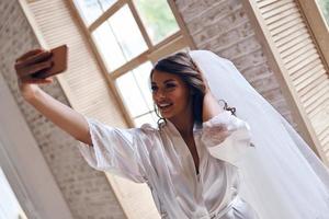 charmante mariée. jolie jeune femme en peignoir de soie et voile souriant tout en prenant selfie près de la fenêtre photo