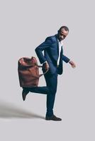 jouer avec style. toute la longueur du beau jeune homme africain en costume complet portant un sac en cuir marron sur son pied en se tenant debout sur fond gris photo