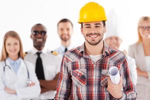 Quand je serai grand, je serai ingénieur. jeune homme confiant dans un casque tenant un plan et souriant tandis qu'un groupe de personnes de différentes professions se tenant à l'arrière-plan photo