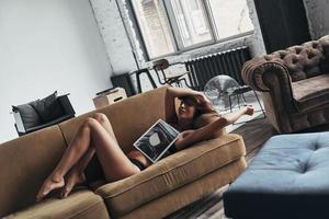 profiter du temps à la maison. jolie jeune femme couverte de magazine souriante allongée sur le canapé à la maison photo
