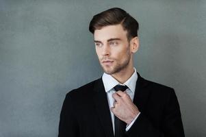 homme d'affaires confiant. confiant jeune homme d'affaires ajustant sa cravate et regardant ailleurs en se tenant debout sur fond gris photo