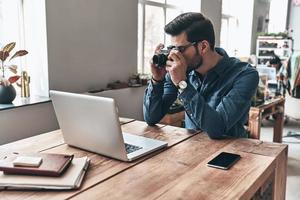 vérification de la caméra. jeune homme réfléchi utilisant un appareil photo tout en travaillant dans l'espace de travail créatif