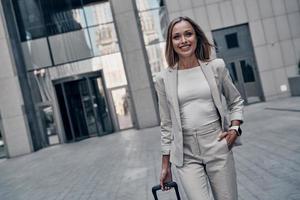 les affaires sont sa vie. belle jeune femme en costume tirant des bagages et souriant tout en marchant à l'extérieur photo