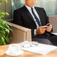 vérification des messages commerciaux. homme mûr confiant en tenues de soirée tapant un message sur son téléphone portable et assis à la chaise à l'extérieur avec une tasse de café au premier plan photo