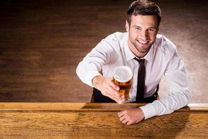 se détendre avec un verre de bière fraîche. vue de dessus du beau jeune homme en chemise et cravate tenant un verre avec de la bière et souriant assis au comptoir du bar photo