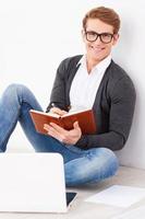 il a un esprit créatif. beau jeune homme à lunettes écrivant quelque chose dans un bloc-notes et souriant assis sur le sol avec un ordinateur portable près de lui photo