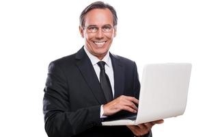 soutenir votre entreprise. homme mûr confiant en tenues de soirée travaillant sur ordinateur portable et souriant tout en se tenant isolé sur fond blanc photo