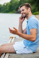 bonne conversation avec un ami. vue latérale d'un beau jeune homme parlant au téléphone portable et souriant assis au bord de la rivière photo