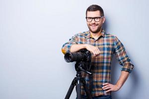 photographe professionnel. portrait d'un jeune homme confiant en chemise tenant la main sur l'appareil photo sur un trépied en se tenant debout sur fond gris