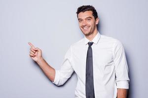 homme d'affaires pointant l'espace de copie. heureux jeune homme en chemise et cravate regardant la caméra et souriant tout en se tenant sur fond gris et pointant l'espace de copie photo