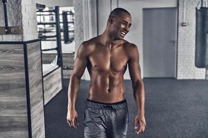 en parfait état. jeune homme africain torse nu regardant loin et souriant tout en se tenant dans la salle de gym photo
