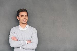 regardant à l'avenir avec le sourire. jeune homme confiant en gardant les bras croisés et en détournant les yeux avec le sourire en se tenant debout sur fond gris photo