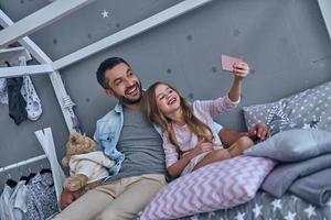 père joyeux et sa petite fille prenant selfie assis sur le lit à la maison photo