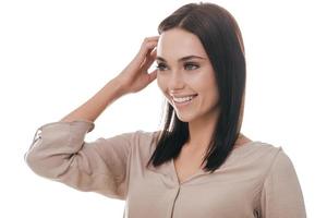 toujours de bonne humeur. séduisante jeune femme tenant la main dans les cheveux et en détournant les yeux avec le sourire en se tenant debout sur fond blanc photo