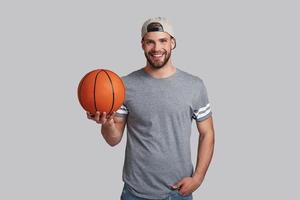 vrai joueur. beau jeune homme souriant portant un ballon de basket et regardant la caméra en se tenant debout sur fond gris photo