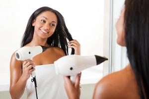 sécher les cheveux dans la salle de bain. vue arrière de la belle jeune femme africaine séchant les cheveux et souriant debout dans la salle de bain et contre un miroir photo