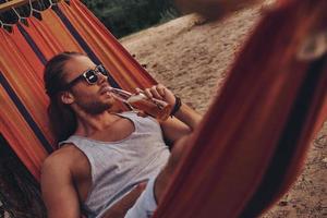 temps de repos. beau jeune homme buvant de la bière et regardant ailleurs en position couchée dans un hamac photo