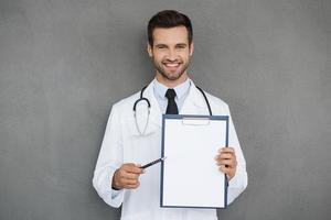 médecin pointant l'espace de copie. souriant jeune médecin en uniforme blanc tenant le presse-papiers et pointant dessus en se tenant debout sur fond gris photo