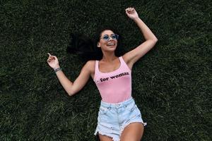 se sentir libre de devenir fou. vue de dessus de jolie jeune femme métisse en maillot de bain et short souriant en position couchée sur l'herbe photo