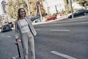 besoin d'un taxi. belle jeune femme en costume saluant un tour et souriant tout en se tenant à l'extérieur photo