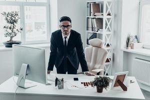 prêt à vous entendre. beau jeune homme en costume complet regardant la caméra avec un visage sérieux tout en s'appuyant sur le bureau photo