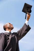 heureux d'être diplômé. vue en angle bas d'un jeune homme africain heureux en robe de graduation tenant son panneau de mortier contre le ciel bleu photo