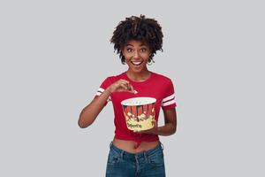 séduisante jeune femme africaine mangeant du pop-corn et regardant la caméra avec le sourire en se tenant debout sur fond gris photo