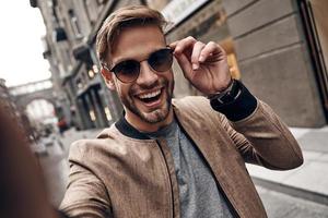 devenir fou. autoportrait d'un beau jeune homme en tenue décontractée souriant et ajustant ses lunettes tout en se tenant à l'extérieur photo