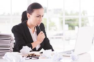 en attente d'inspiration. jeune femme réfléchie en tenues de soirée sur un ordinateur portable et touchant son menton avec un stylo alors qu'elle était assise sur son lieu de travail photo