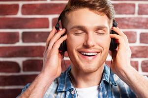 appréciant la musique. beau jeune homme écoutant le lecteur mp3 et gardant les yeux fermés tout en se tenant contre le mur de briques photo