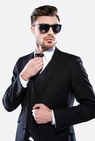 élégant et charmant. portrait de beau jeune homme en tenues de soirée et lunettes de soleil ajustant sa cravate en se tenant debout sur fond gris photo