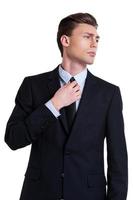 confiant et réussi. jeune homme confiant en tenues de soirée ajustant sa cravate et regardant ailleurs tout en se tenant isolé sur blanc photo