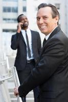 il est le meilleur dans son domaine. homme âgé joyeux en tenues de soirée se déplaçant par des escaliers et regardant par-dessus l'épaule tandis qu'un homme africain parlant au téléphone mobile en arrière-plan photo