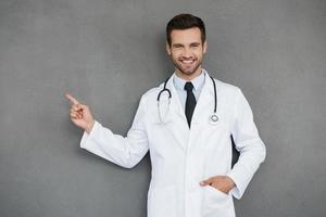 montrant de nouvelles façons de guérir. souriant jeune médecin en uniforme blanc regardant la caméra et pointant vers l'extérieur en se tenant debout sur fond gris photo