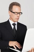 homme d'affaires avec ordinateur portable. homme mûr confiant en tenues de soirée travaillant sur ordinateur portable en se tenant debout sur fond gris photo